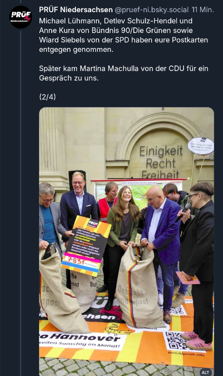 Zu sehen sind Menschen aus dem Landtag, die die Übergroße Postkarte entgegennehmen. Auf der Postkarte steht geschrieben, dass 7831 Mal Postkarten eingereicht sind. Die Menschen wirken glücklich. Vor sich stehend haben sie 3 große Postsäcke, in denen sie teilweise interessiert hineinschauen. Im Hintergrund gibt es etwas verdeckt eine Landkarte von Niedersachsen. Dahinter ist der Niedersächsische Landtag mit der Aufschrift "Einigkeit, Recht, Freiheit".